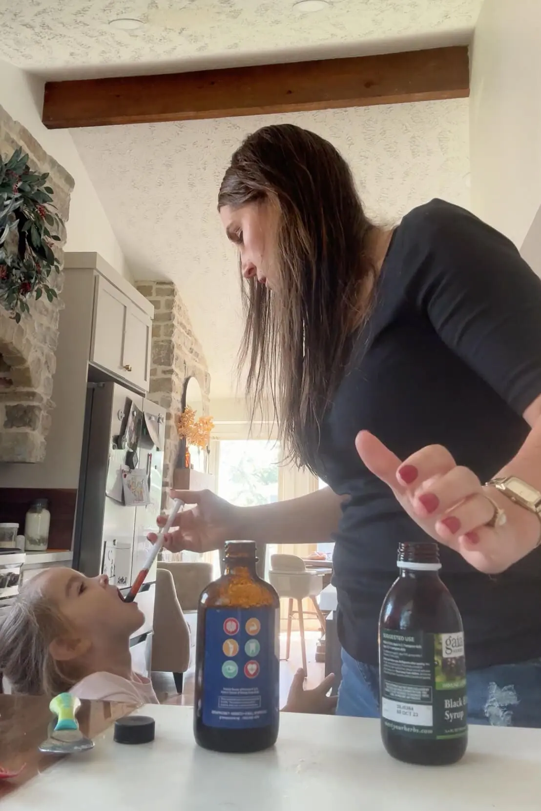 Mother giving a young child a liquid cod liver oil supplement using a syringe in a bright kitchen setting.